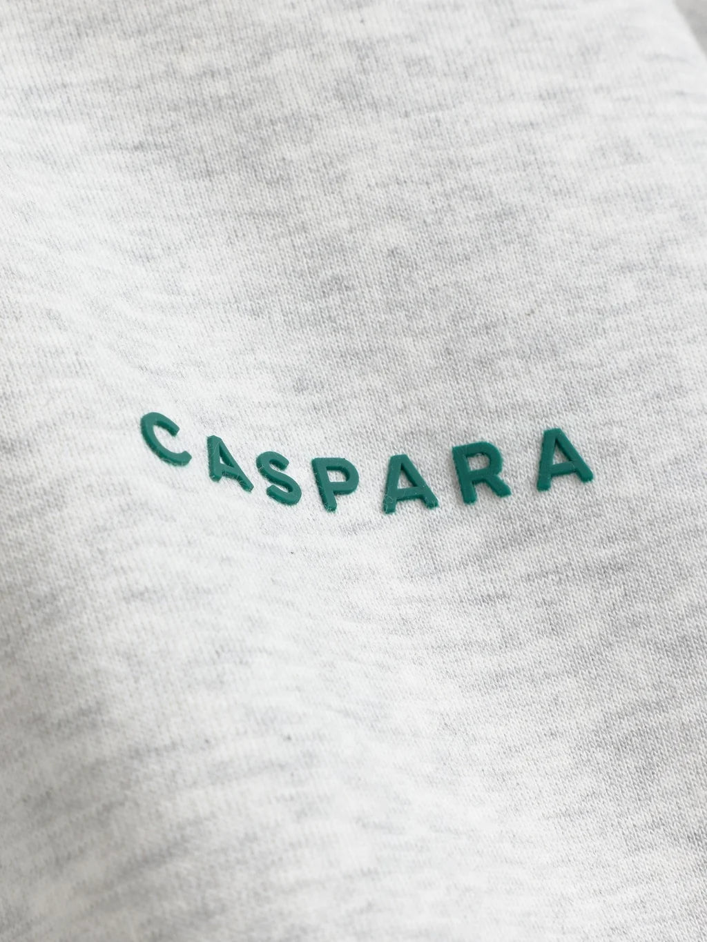 Close-up detail of the green CASPARA logo on grey melange sweatshirt fabric.